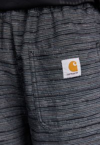 Carhartt šorts mrke boje sa teksturiranim, prugastim uzorkom. Imaju stražnji džep i kvadratnu oznaku s žutim emblemom.