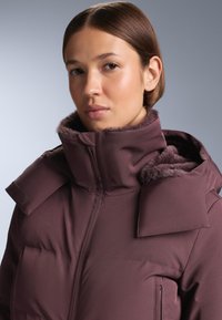 Gewatteerde jas in donker mauve met hoge kraag, nepbontvoering en ritszakken. Heeft oversized schouderaccenten en een gladde buitenkant.