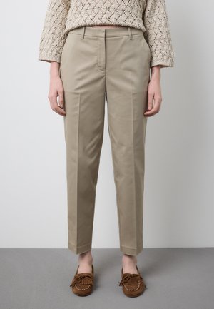 Person trägt beige maßgeschneiderte Hosen, einen cremefarbenen Strickpullover und braune Mokassin-Schuhe, steht auf einem grauen Boden vor einer weißen Wand.