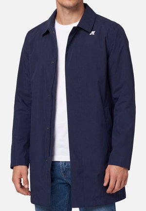 Uomo che indossa un cappotto lungo blu scuro sopra una maglietta bianca e jeans blu, in piedi con le braccia rilassate lungo i fianchi.