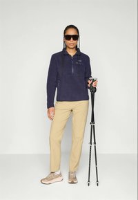 Navy fleece pullover with a quarter zip, tan pants, and beige hiking shoes. Model holds trekking poles and wears dark sunglasses.
