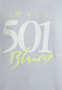 Lys grå stof med store hvide bogstaver "Levi's 501" og mindre gule kursiv "Blues" på en glat, let struktureret overflade.