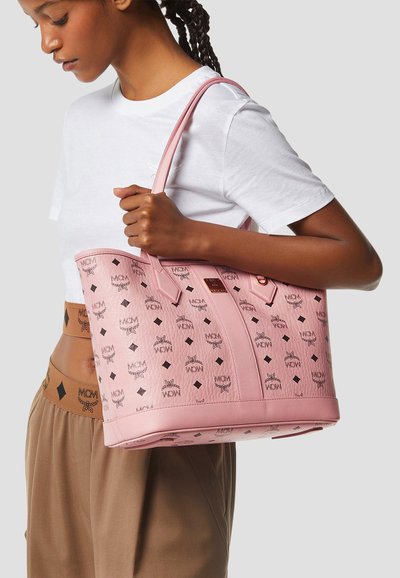 Donna con t-shirt bianca e pantaloni marroni tiene una grande borsa tote rosa MCM con motivo logo sulla spalla su sfondo semplice.
