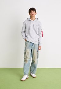 Felpa grigia chiara con tasca frontale e logo, abbinata a jeans cargo blu chiaro con tasche laterali e una texture sbiadita. Sneakers bianche.
