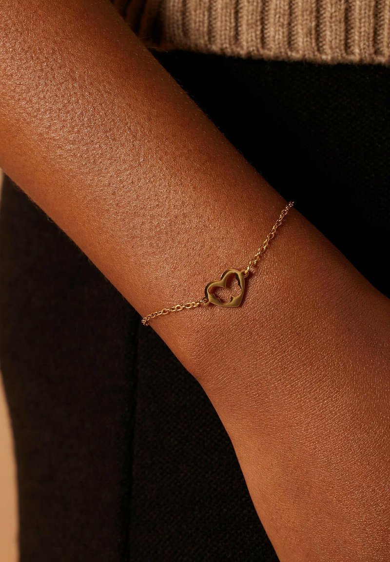 Gouden kettingarmband met een hartvormige bedel met een open midden, rustend op een gestructureerd huidoppervlak.