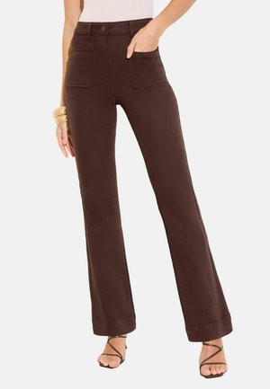 Naine, kes kannab kõrge vöökohaga tumepruune sirgeid pükse esiseintega taskutega ja mustade rihmadega kontsakingi.