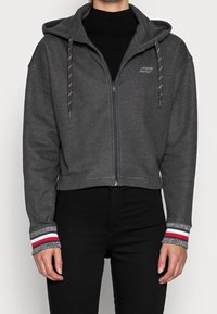 Sudadera gris con cremallera y diseño corto, que presenta una capucha con cordones y puños de canalé con acentos en rojo, blanco y gris. Textura suave.