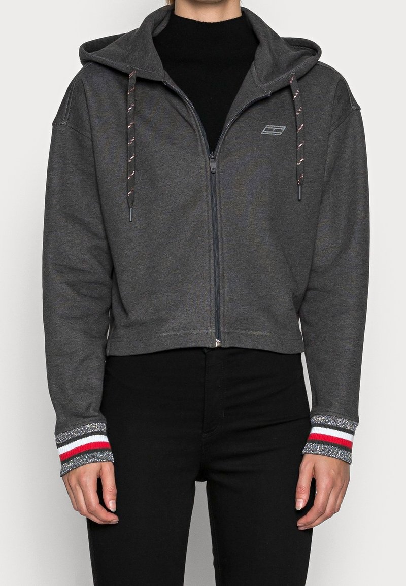 Sudadera gris con cremallera y diseño corto, que presenta una capucha con cordones y puños de canalé con acentos en rojo, blanco y gris. Textura suave.
