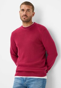 Maglione rosso a maglia con texture a coste, scollatura a girocollo e spacco laterale. Abbinato a jeans azzurri chiari per un look casual.
