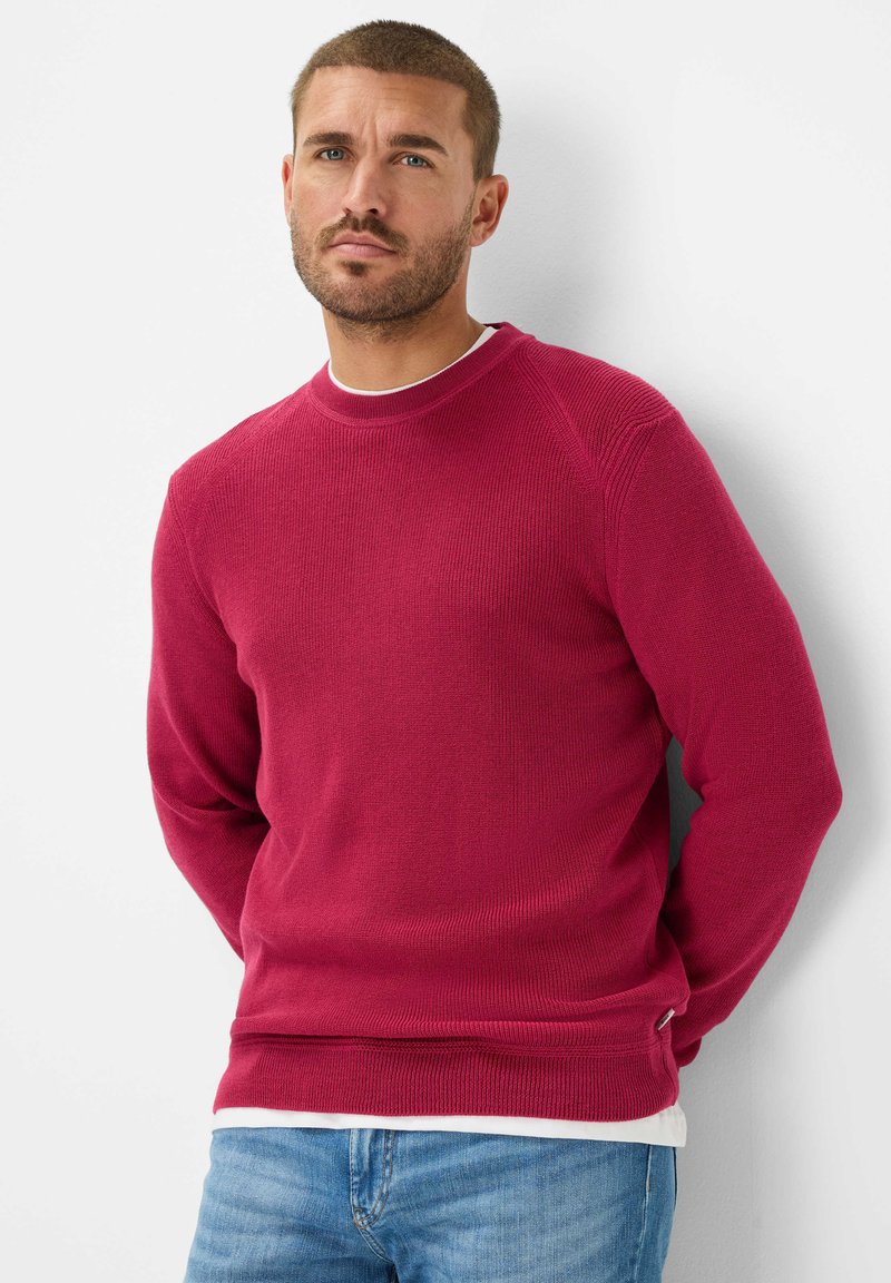 Maglione rosso a maglia con texture a coste, scollatura a girocollo e spacco laterale. Abbinato a jeans azzurri chiari per un look casual.
