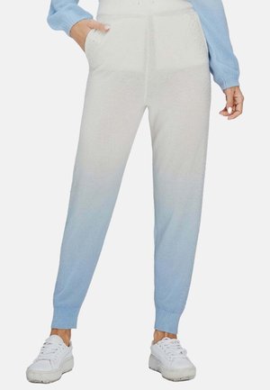Frau trägt hellblaue Ombre-Strick-Jogginghose, blauen Pullover und weiße Sneakers, steht mit einer Hand in der Tasche.
