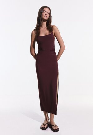 Žena stojí so založenými rukami za chrbtom, oblečená v bezrukávových bordových maxi šatách s bočným rozparkom a čiernymi sandálmi na platforme na bielom pozadí.