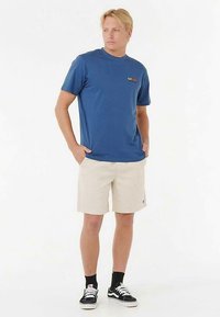 Camiseta de algodón azul con un logo colorido, combinada con pantalones cortos beige y zapatillas negras. El conjunto presenta un ajuste casual y mangas cortas.