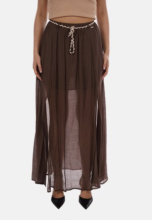 Donna indossa una lunga gonna marrone trasparente con vita elasticizzata e una cintura beige intrecciata, abbinata a scarpe nere con punta appuntita e un top corto.