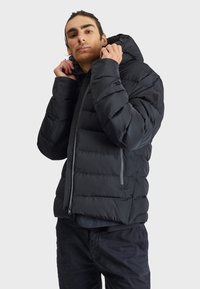 Black puffer jacket with a zip-up front, textured fabric, and adjustable hood. Features a small logo on the left shoulder and side pockets.