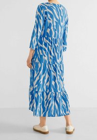Lång blå klänning med ett vågigt vitt mönster, trekvartsärmar, lagerdesign och rundad halsringning, i kombination med vita sneakers.