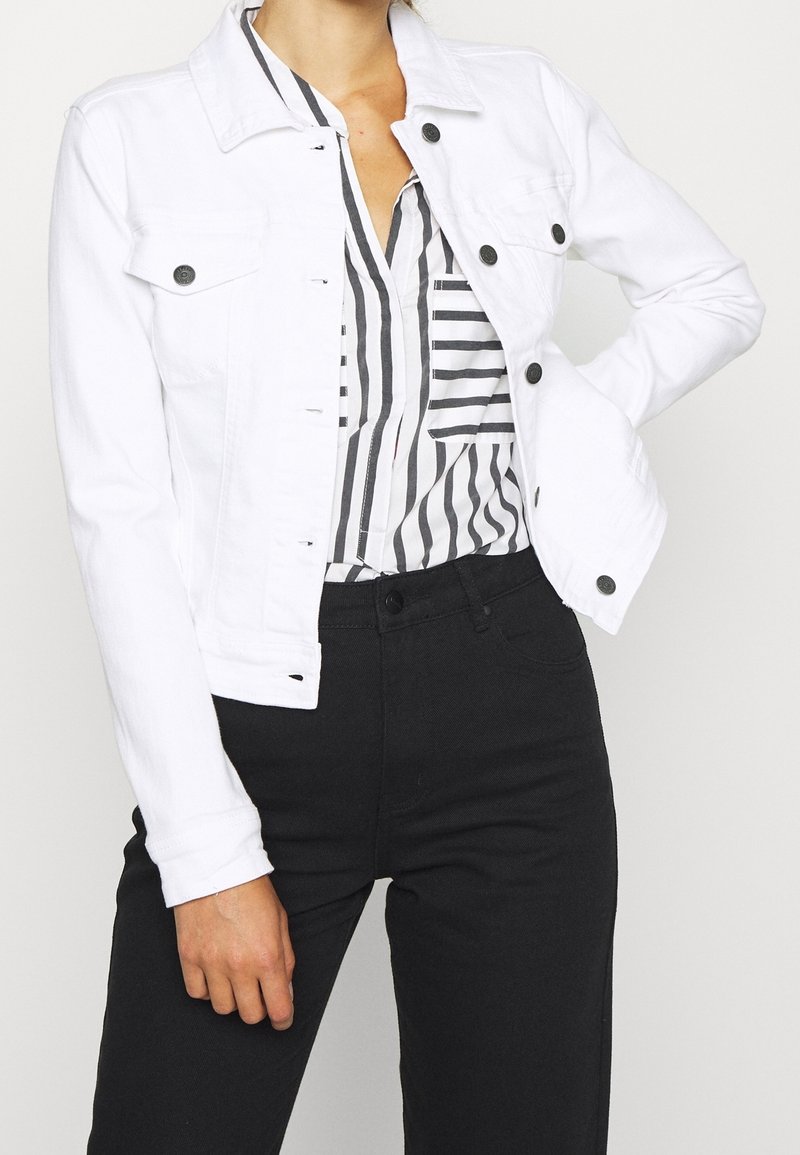 Veste en denim blanche à manches longues, boutons en métal noir et poches avant. Associée à un chemisier rayé et un pantalon noir.