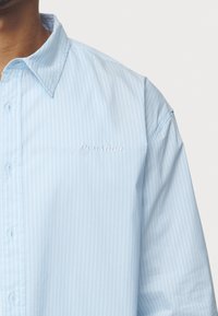 Svetlo modrá košeľa s krátkym rukávom a vertikálnymi bielymi pruhmi, klasickým golierom a vyšívaným logom "PEGADOR" na hrudi.