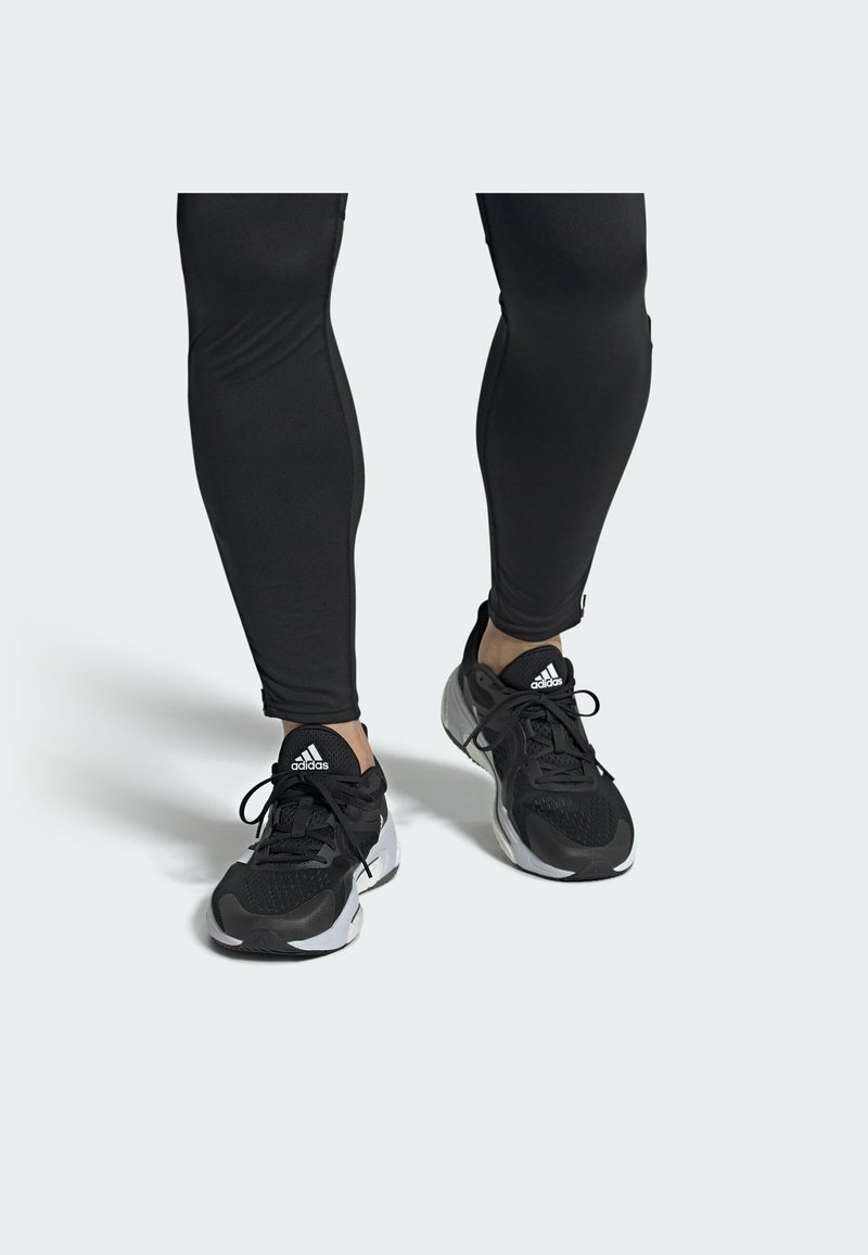 Schwarze Sportschuhe mit strukturiertem Obermaterial, weißen Akzenten und schwarzen Schnürsenkeln, kombiniert mit enganliegenden schwarzen Leggings. Statischer Hintergrund.