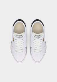 Ζευγάρι λευκών αθλητικών παπουτσιών Reebok με χρυσό λογότυπο, λεπτομέρεια μαύρης φτέρνας και λευκά κορδόνια, εμφανιζόμενα από πάνω σε απλό φόντο.