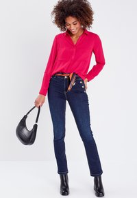 Chemise à manches longues rose vif avec des détails de boutons, associée à un jean taille haute en denim foncé avec des boutons dorés et des bottines noires.