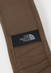 Brown padded strap with a textured surface, featuring a black fabric label displaying "THE NORTH FACE" in gray lettering.