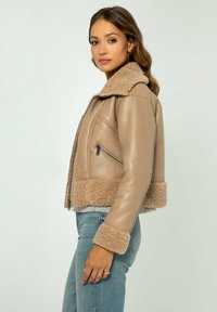 Giacca in pelle marrone foderata in shearling con un design corto, tasche con zip e polsini texturizzati. Abbinata a jeans di denim blu chiaro.