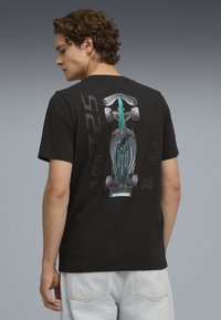 Camiseta de algodón negra con un gráfico de un coche de carreras verde en la parte posterior, con "AMR25" y "2025" impresos en blanco y acentos metálicos.