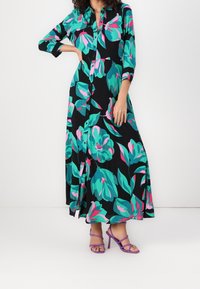 Robe longue noire avec un grand motif floral vert et rose, devant à boutons, manches trois-quarts ; associée à des talons à brides violets.