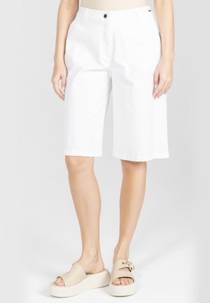 Kobieta ubrana w białe krótkie spodenki do kolan oraz beżowe sandały na platformie, stojąca na tle jasnego, jednolitego tła.