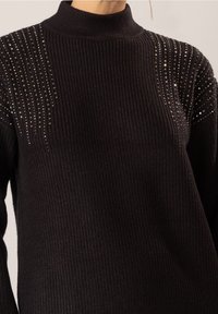 Pull noir en tricot avec un col montant, doté d'une texture côtelée et d'ornements en cristaux décoratifs le long des épaules formant un motif ondulé.