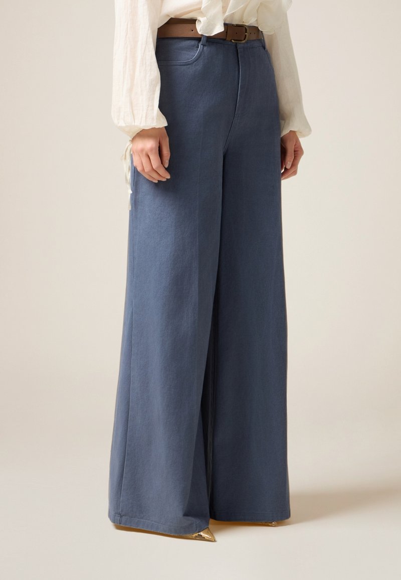 Weite Denim-Hosen in Hellblau, mit hohem Bund und subtiler Textur. Kombiniert mit einem braunen Gürtel, ergänzt durch ein beiges Oberteil.