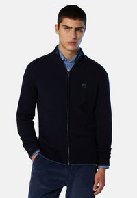 Maglione blu navy con cerniera, polsini e collo a costine, realizzato in morbido tessuto a maglia. Presenta un logo circolare sul lato sinistro del petto.