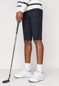 Bermudas azul-marinho com uma textura suave, combinadas com sapatilhas brancas e detalhes cinzentos. O sujeito segura um taco de golfe.