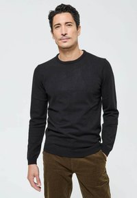 Pull en tricot noir avec un col rond, des manches longues et des bords riboutés. Porté avec un pantalon en velours côtelé beige. Texture lisse et design ajusté.