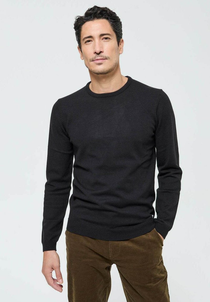 Pull en tricot noir avec un col rond, des manches longues et des bords riboutés. Porté avec un pantalon en velours côtelé beige. Texture lisse et design ajusté.
