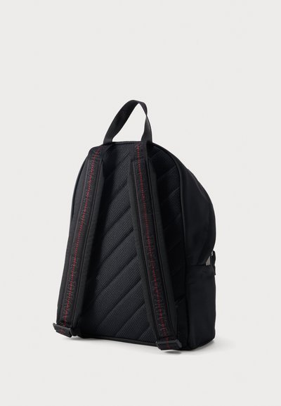 Sac à dos noir avec bretelles rembourrées ornées de surpiqûres rouges et une poignée supérieure, vu de dos sur un fond clair uni.