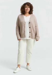 Beige ribstrikket cardigan med store knapper, båret over en hvid skjorte. Bukserne er cremefarvede. Modellen har krøllet hår og hvide sneakers.