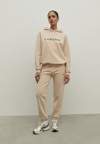 Béžová súprava s kapucňou a teplákmi s logom "LAMANIA" v čiernej farbe, textúrovaná tkanina, rebrované manžety a sťahovacia šnúrka v páse. V kombinácii so striebornými teniskami.