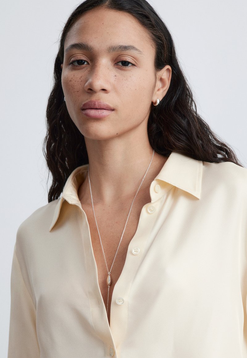 Jeune femme aux longs cheveux foncés et taches de rousseur, portant une chemise crème boutonnée, de petites boucles d'oreilles en créole et un collier pendentif en argent.