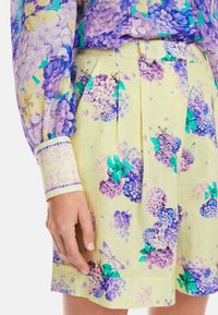 Gelbe Shorts mit floralem Druck, der lila Hortensien und grüne Blätter zeigt. Falten an der Taille und ein dekorativer Saumakzent.