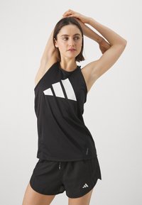 Černý bezramínkový sportovní top s bílým geometrickým logem, doplněný černými síťovanými šortkami s bílým logem značky, lehký materiál.