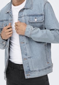 Ljusblå denimjacka med klassisk skärning, knappstängning, två framfickor och en något bleknad textur. Bärs över en vit skjorta.