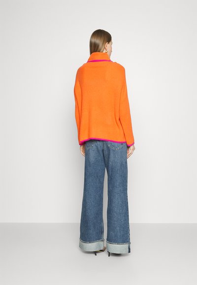 Oversized orange knit sweater with a ribbed texture, featuring a folded collar and pink accent at the hem, paired with wide-leg denim jeans.