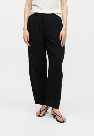 Žena nosiaca voľné čierne široké nohavice a čierne sandále na podpätku s tenkými remienkami, stojaca pred jednoduchým svetlým pozadím.