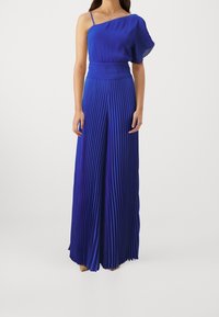 Szafirowy, jednoskrzydłowy, plisowany kombinezon z dopasowaną talią i zwiewnym krojem. Posiada błyszczący, teksturowany materiał oraz cienki ramiączko.