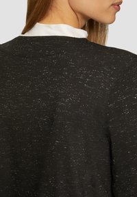 Schwarzes Strickoberteil mit strukturierter Oberfläche, das subtile metallic Fäden aufweist, einem runden Halsausschnitt und einem entspannten Schnittdesign. Akzente sind auf dem Stoff sichtbar.