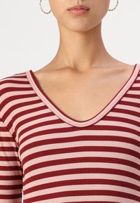 Rot- und rosa gestreiftes Oberteil mit geripptem Material, das einen tiefen V-Ausschnitt und lange Ärmel hat. Runde Creolen sind sichtbar.