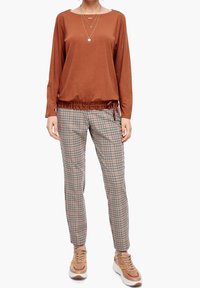 Długi rękaw, rdzawy top z marszczonym dołem, w połączeniu z spodniami w kratkę w kolorach białym, brązowym i czarnym. Noszony z jasnobeżowymi i pomarańczowymi sneakersami.