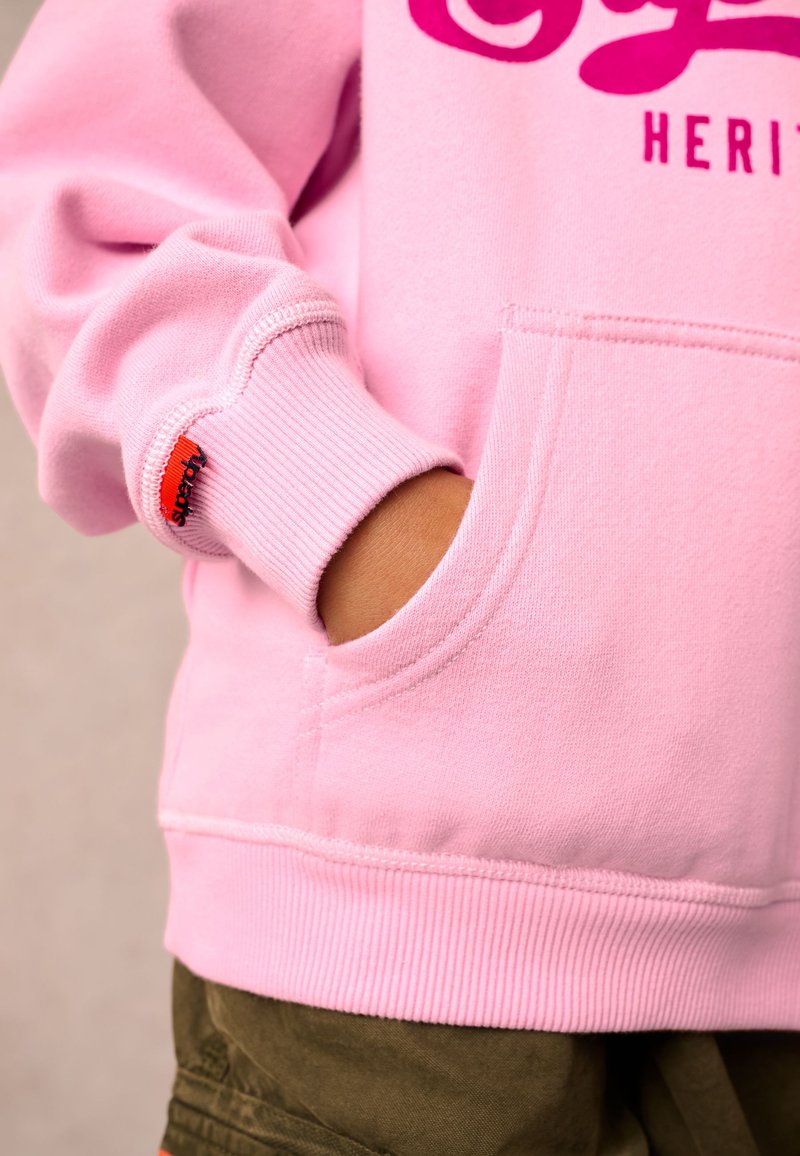 Felpa rosa chiaro con tasca frontale, polsini a costine e logo in rosa più scuro. Presenta un'etichetta brandizzata rossa sulla manica. Tessuto morbido.